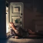 A woman relaxes near an open refrigerator in a dimly lit room, creating a serene and moody atmosphere.
