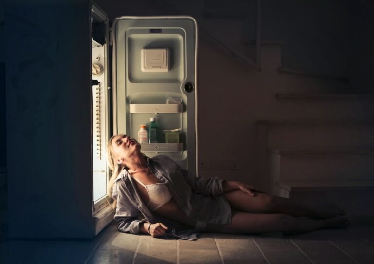 A woman relaxes near an open refrigerator in a dimly lit room, creating a serene and moody atmosphere.