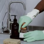 Hands in latex gloves washing dishes with soap at a kitchen sink.