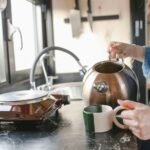 A woman in a denim jacket pours tea from a kettle into a cup on a kitchen counter.