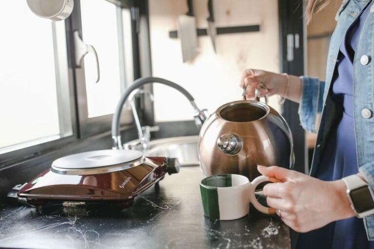A woman in a denim jacket pours tea from a kettle into a cup on a kitchen counter.