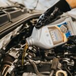 A mechanic wearing gloves pouring oil into a car engine in a garage setting.