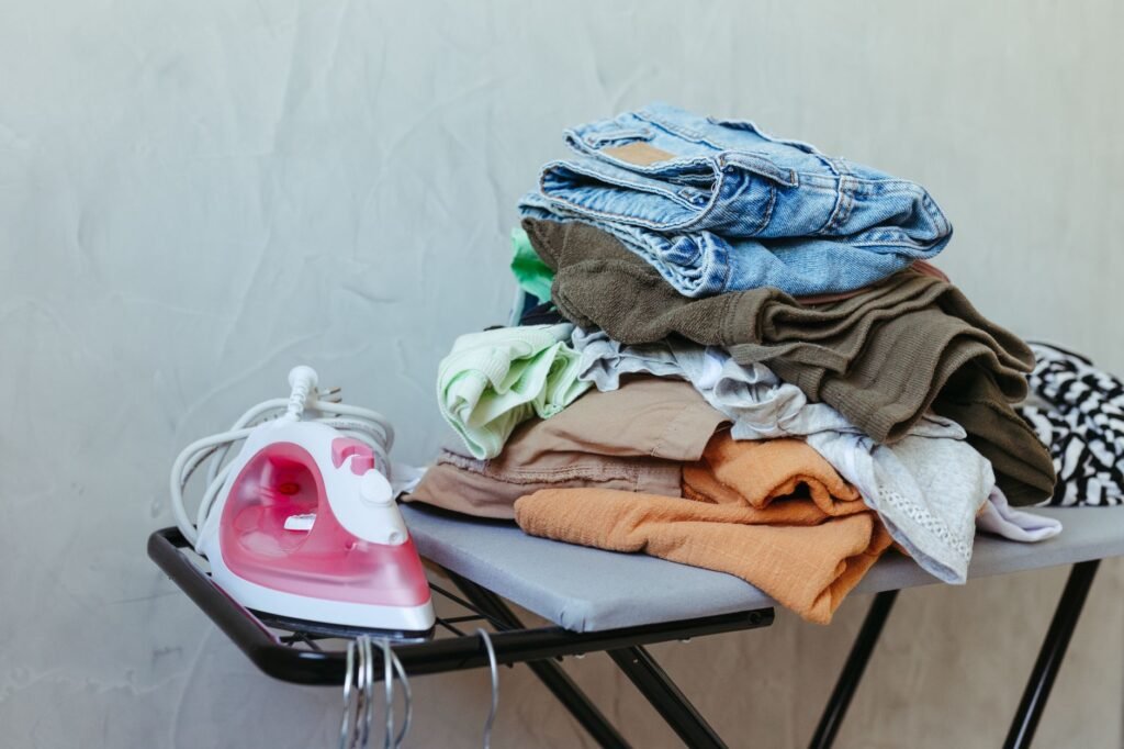 A stack of freshly ironed clothes next to a steam iron on an ironing board, ready for organizing.