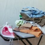 A stack of freshly ironed clothes next to a steam iron on an ironing board, ready for organizing.