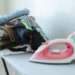 Tidy stack of clothes ready to be ironed with an iron on an ironing board.