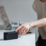 Close-up of a hand reaching for a wireless speaker on a desk beside a laptop.