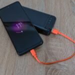 A smartphone charging from a power bank with an orange cable on a wooden table.