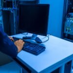 An IT professional operates a computer in a server room, managing network systems and connected devices.