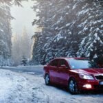 A red car drives along a snowy forest road in winter, surrounded by frosty trees.