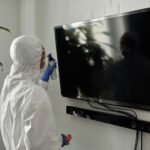 A person wearing PPE disinfecting a TV screen inside a room during the COVID-19 pandemic.