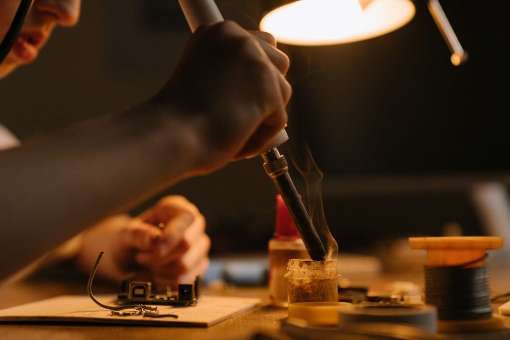 Focused close-up of a person soldering a printed circuit board under a desk lamp.
