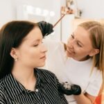 Makeup artist applying eyebrow tint on a smiling woman in a beauty salon.