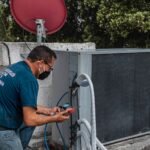 Technician repairing an air conditioner unit outdoors, wearing a facemask and using a manifold gauge.