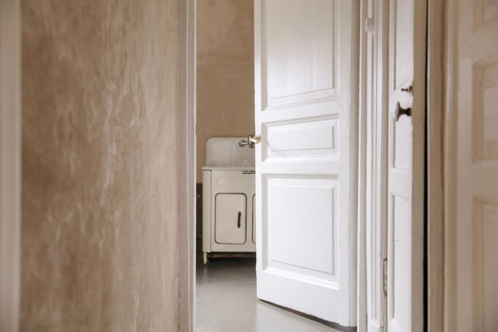 A rustic interior with a white wooden door slightly ajar revealing a vintage sink inside.