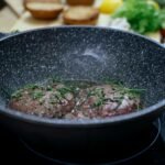 Juicy cutlets with rosemary frying in pan on black stove in kitchen with various ingredients for burger on blurred background