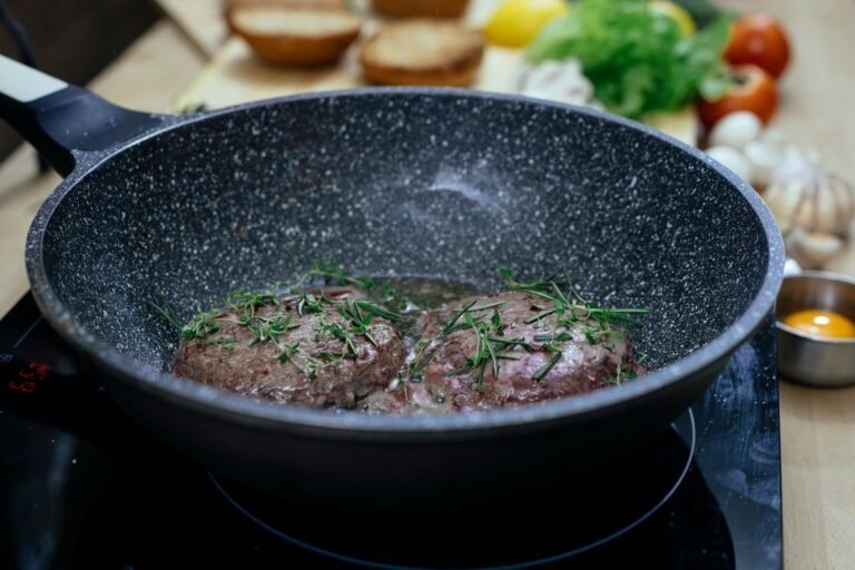 Juicy cutlets with rosemary frying in pan on black stove in kitchen with various ingredients for burger on blurred background