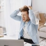 An upset man with mouth open, pulling hair at a desk with a laptop in a home setting.