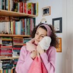Young woman hugging blankets in a cozy room surrounded by bookshelves, expressing warmth.