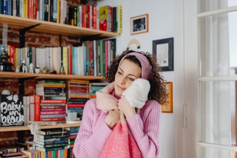 Young woman hugging blankets in a cozy room surrounded by bookshelves, expressing warmth.