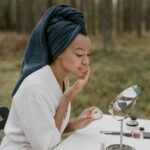 A woman in a bathrobe applies face cream in a serene outdoor environment.