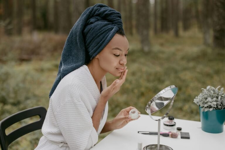 A woman in a bathrobe applies face cream in a serene outdoor environment.