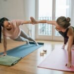 Two women performing yoga plank exercises on mats with a supportive high-five indoors.