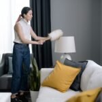 Asian woman dusting a lampshade in a modern living room with yellow pillows.