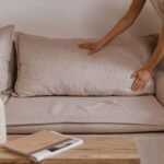 Minimalistic living room scene with a person arranging a pillow on a beige sofa.