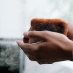 A close-up view of hands holding a toasted slice of bread with visible smoke and burnt texture.
