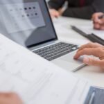 Close-up of hands working on documents and a laptop in an office setting, illustrating teamwork and productivity.