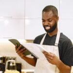 A chef wearing an apron looks puzzled while reading a recipe in a contemporary kitchen.