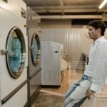 A man sits in a laundromat next to industrial washing machines, waiting for his laundry.