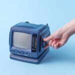 A hand is adjusting a vintage portable TV set against a blue background, highlighting retro technology.
