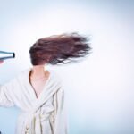 Woman in a bathrobe using a hairdryer, hair flying wildly. Indoor setting.