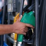 Close-up of a hand holding a green fuel nozzle at a gas pump station outdoors.