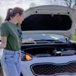 A woman looks puzzled by her broken car with an open hood on a rural road.