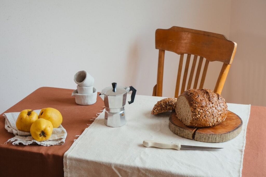 A warm kitchen scene with sliced bread, fruits, and a stovetop coffee maker on a rustic table.