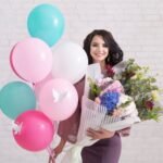 Young woman with balloons and floral bouquet celebrates indoors with a smile.