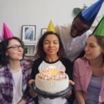 Group of friends celebrating a birthday with a cake and party hats indoors.