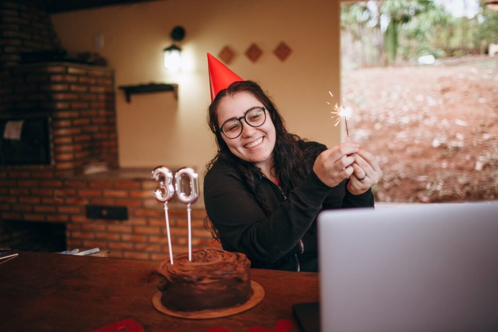 Cheerful woman in party hat holding sparkler with chocolate cake for 30th birthday celebration indoors.