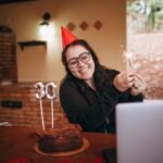 Cheerful woman in party hat holding sparkler with chocolate cake for 30th birthday celebration indoors.
