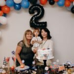A family celebrates a child's second birthday with balloons and a themed cake.