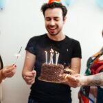 Three friends celebrate a birthday with cake and sparklers indoors wearing party hats.