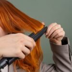 Close-up of a woman styling her red hair using a hair straightener against a green background.