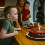 Young boy celebrating birthday by blowing candles on chocolate cake with raspberries.
