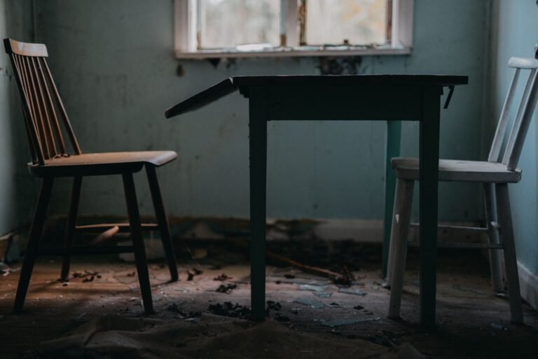 Moody interior of an abandoned dining room featuring wooden chairs and a table.