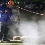 Man in protective gear using a high-pressure washer amid steam at night in an industrial setting.
