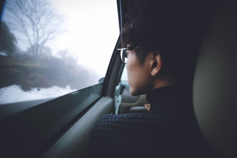 Side view of a thoughtful person with eyewear gazing out of a car window into a foggy winter landscape.