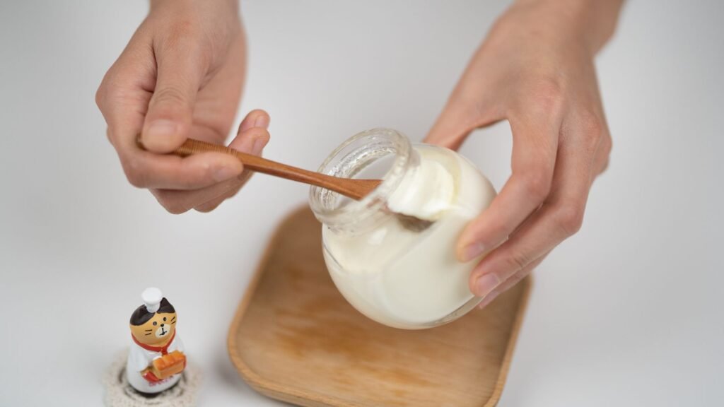 From above crop faceless male eating organic white yogurt from glass jar by using bamboo spoon