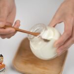 From above crop faceless male eating organic white yogurt from glass jar by using bamboo spoon